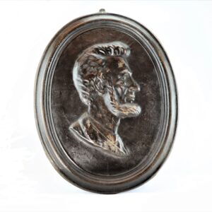 Oval relief plaque depicting Abraham Lincoln's profile with distinct facial features and a beard, set in a dark raised frame against a plain background.