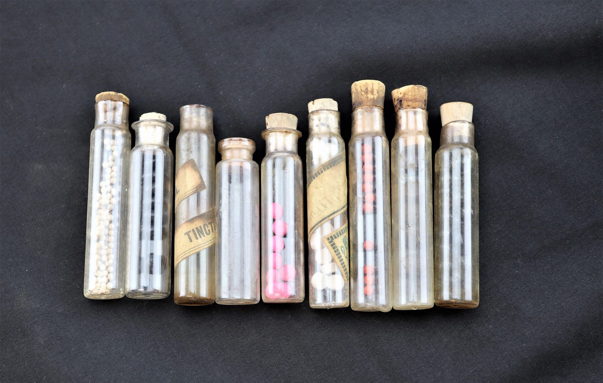 A Leather Apothecary Kit (sold) featuring eight vintage glass vials with cork stoppers—some filled with pills or beads—is displayed side by side against a black background.