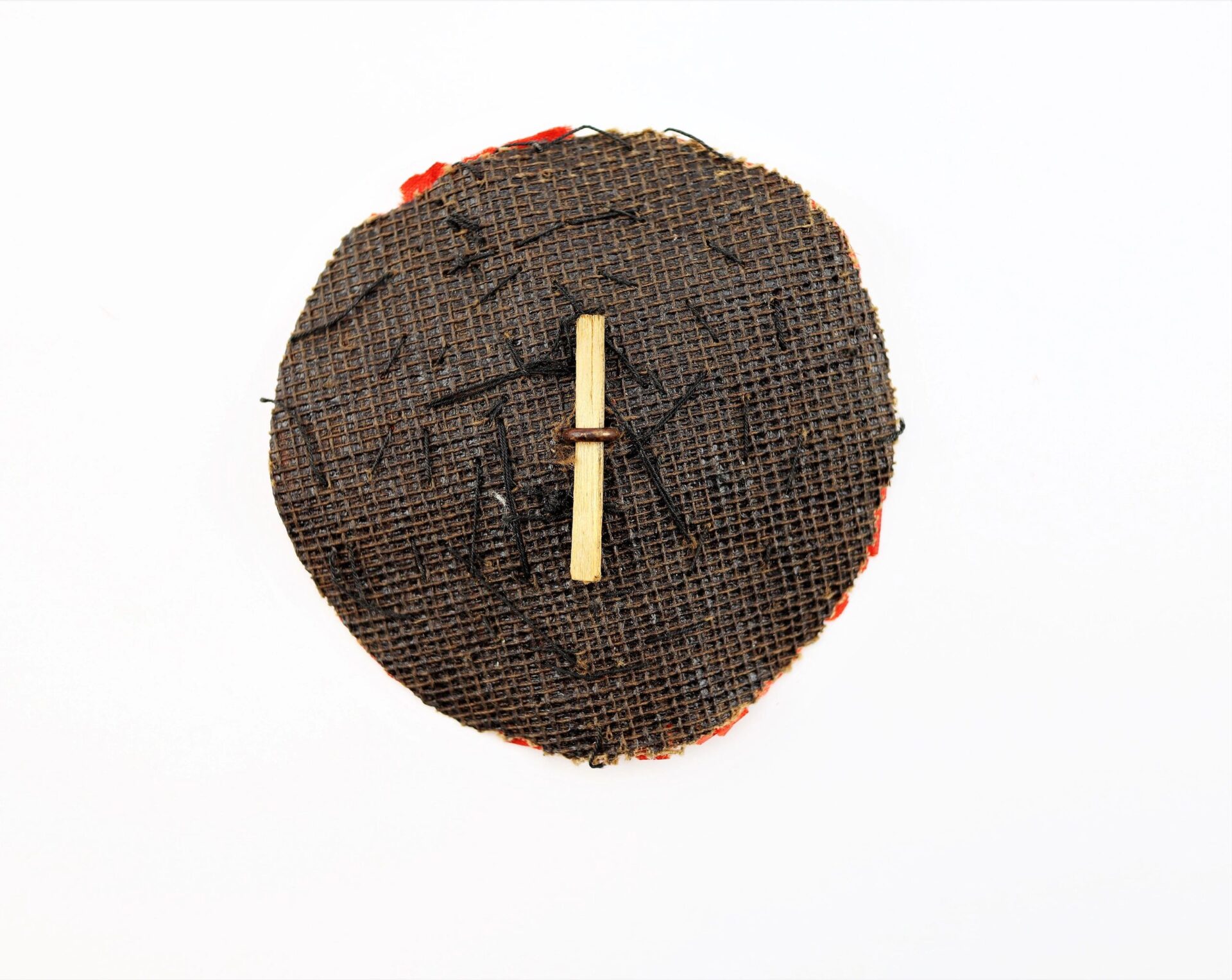 A round, textured dark brown mesh fabric forms the Patriotic Cockade (SOLD), featuring a small beige stick tied vertically at its center, displayed on a plain white background.
