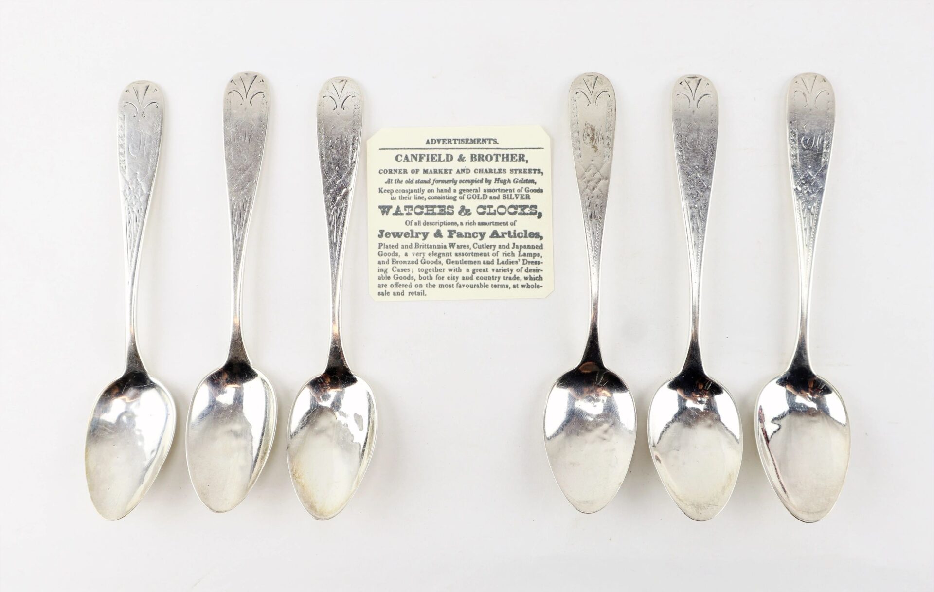 Six Civil War Silver Teaspoons, now sold, are arranged in a row on a white surface with a vintage Canfield & Brother advertisement card placed above them.