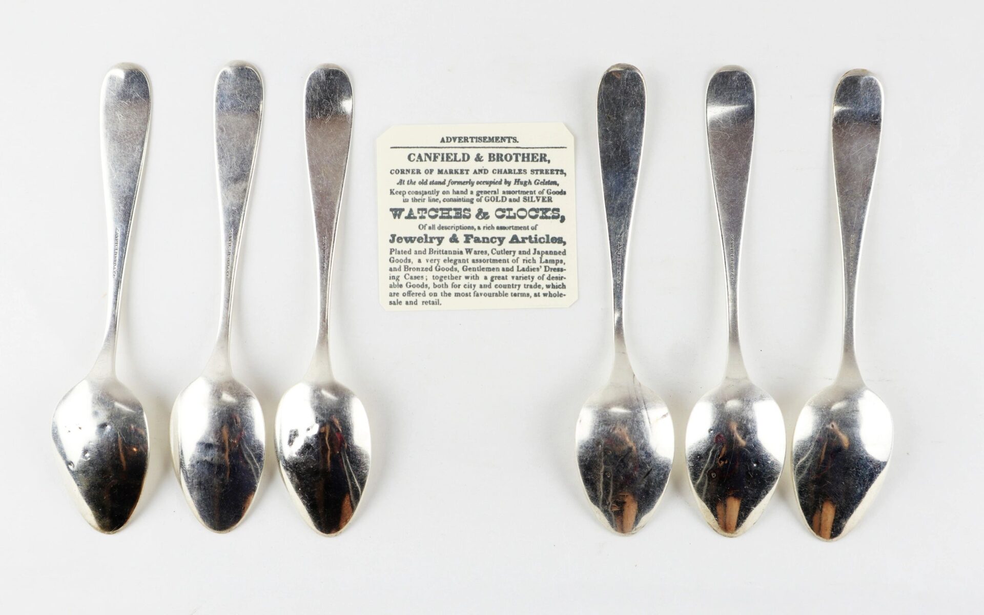 Six Civil War silver teaspoons are arranged in a row around a vintage advertisement card for Canfield & Brother jewelers.