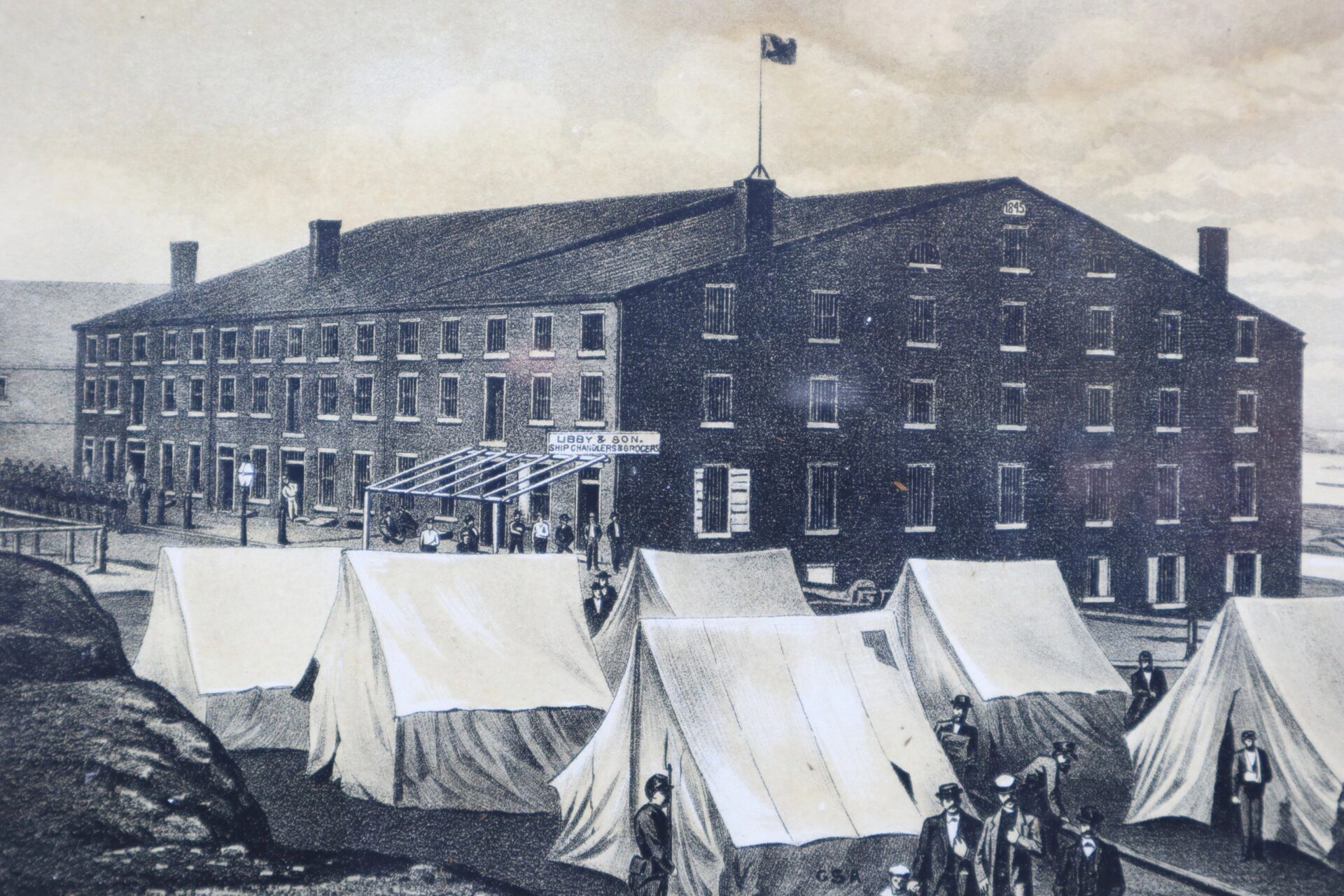 Black-and-white historic illustration of Libby Prison, with a "Sold" sign, tents in front, and people gathered around the large brick building.