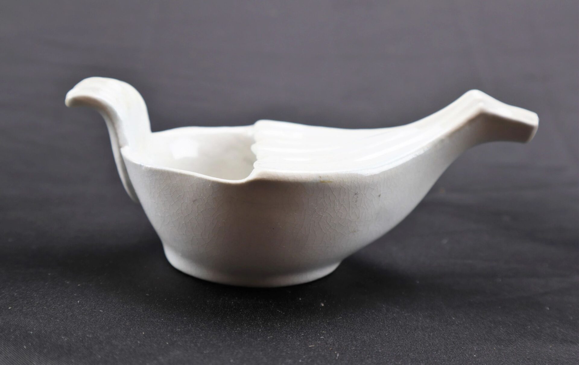 The Invalid Feeder, a white ceramic bowl shaped like a bird with a tail-like handle and beak-shaped spout, is shown against a plain dark background.