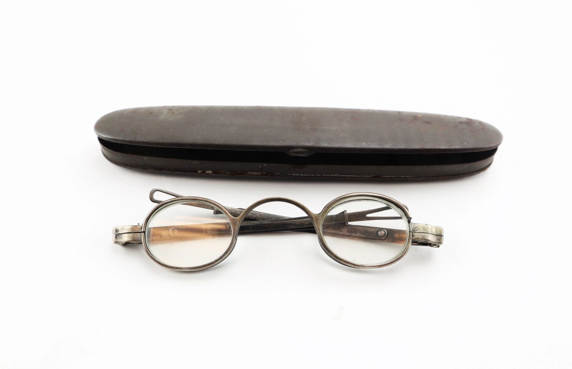 A pair of Civil War Eyeglasses with metal frames and folding arms are displayed in front of a closed, oval metal case on a white background.