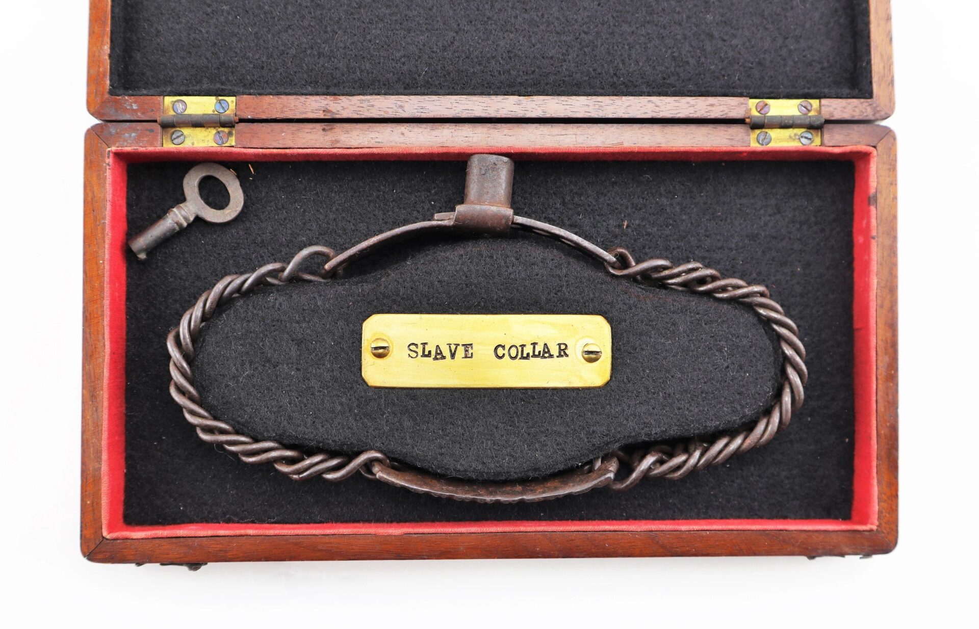 A Civil War dog/slave collar labeled "SLAVE COLLAR," made of iron with brass tag, is displayed in an open wooden box along with a key beside it.