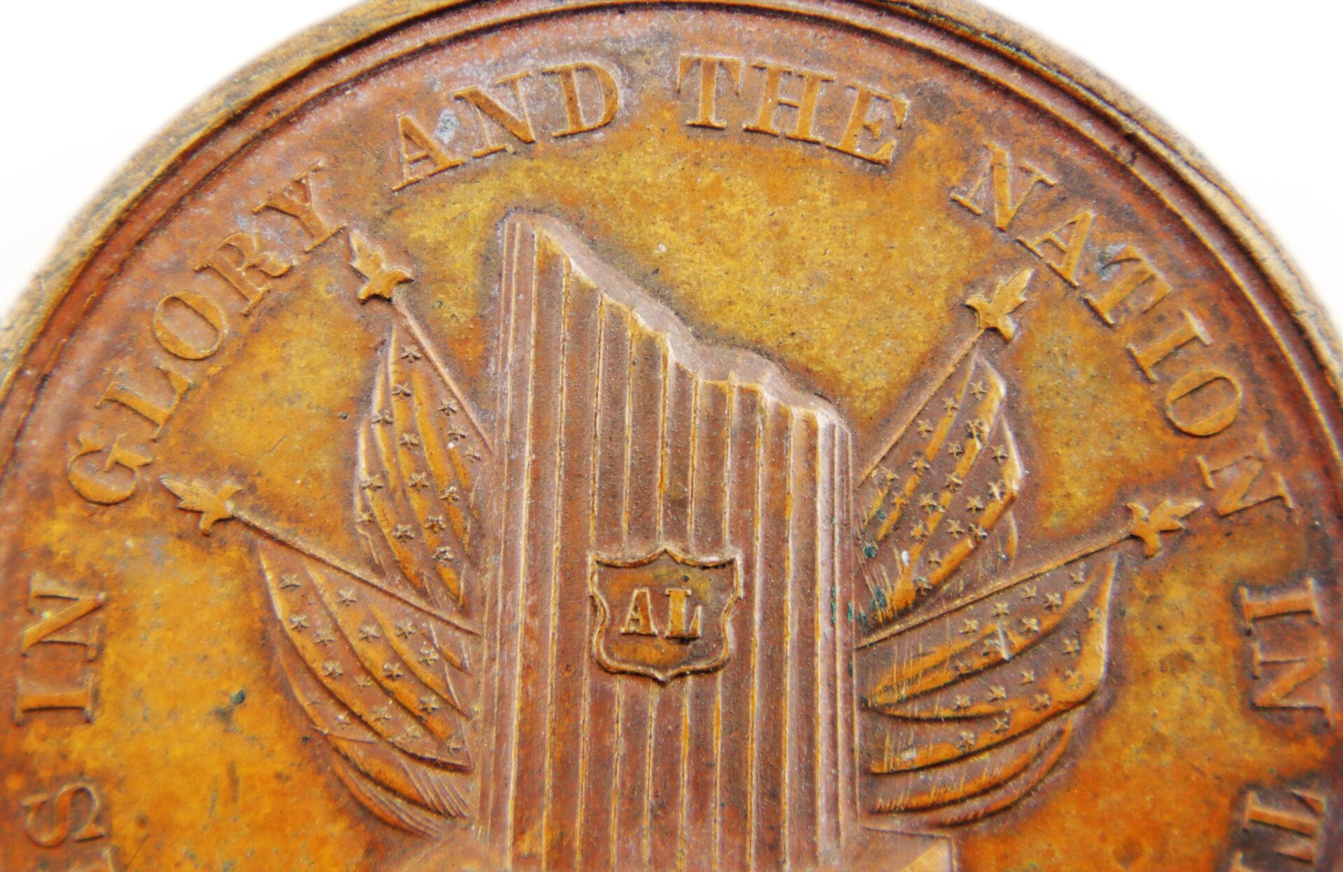 Close-up of a worn Lincoln 1865 Lincoln Assassination Medal (SOLD), displaying part of its inscription, a shield with "AL," and background flags adorned with stars.