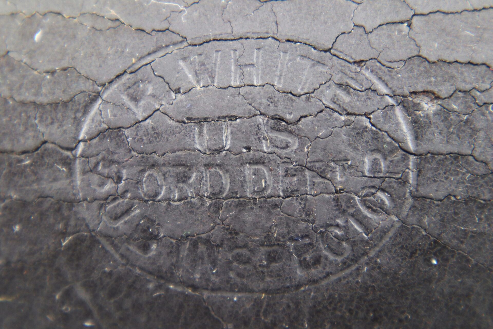 Close-up of a worn, cracked black surface from a Civil War Percussion Cap Box, showing an embossed oval stamp with "U.S. SOLDIER" and other partially legible text.