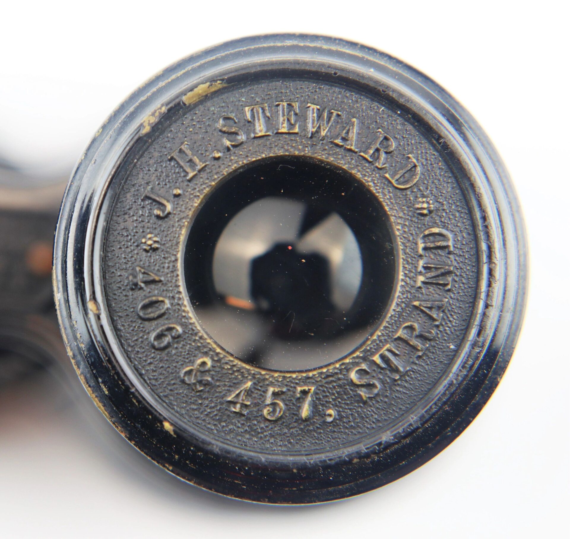 Close-up of Civil War Field Glasses and Case / SOLD, featuring a black optical lens with "J.H. Steward Ltd, 406 & 457, Strand" engraved in metallic text around the edge.