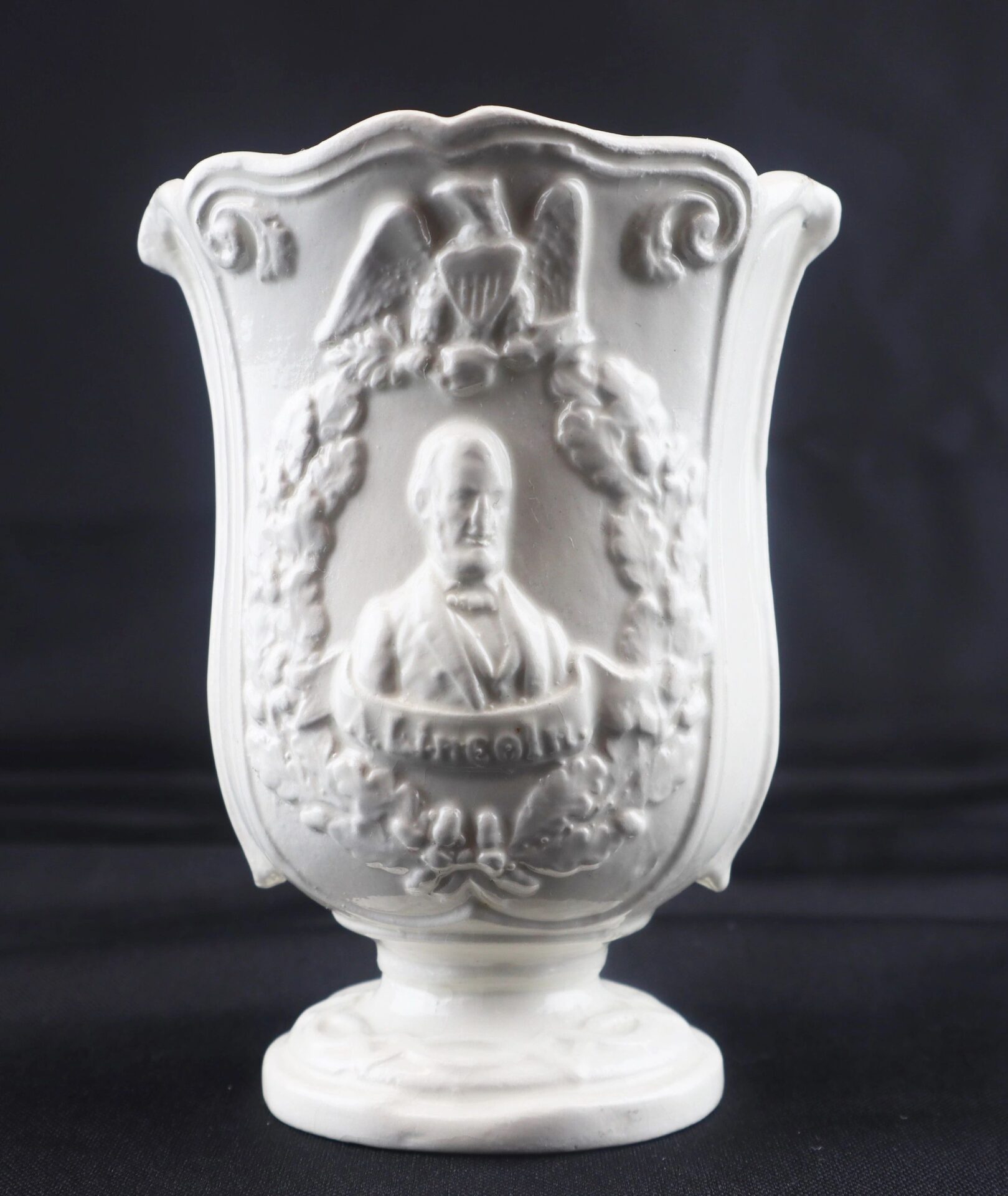 The Civil War Vintage Abraham Lincoln Stoneware Vase is a white ceramic piece with a raised relief of Lincoln, an eagle, a wreath, and "Lincoln" on the front, all set against a black background.
