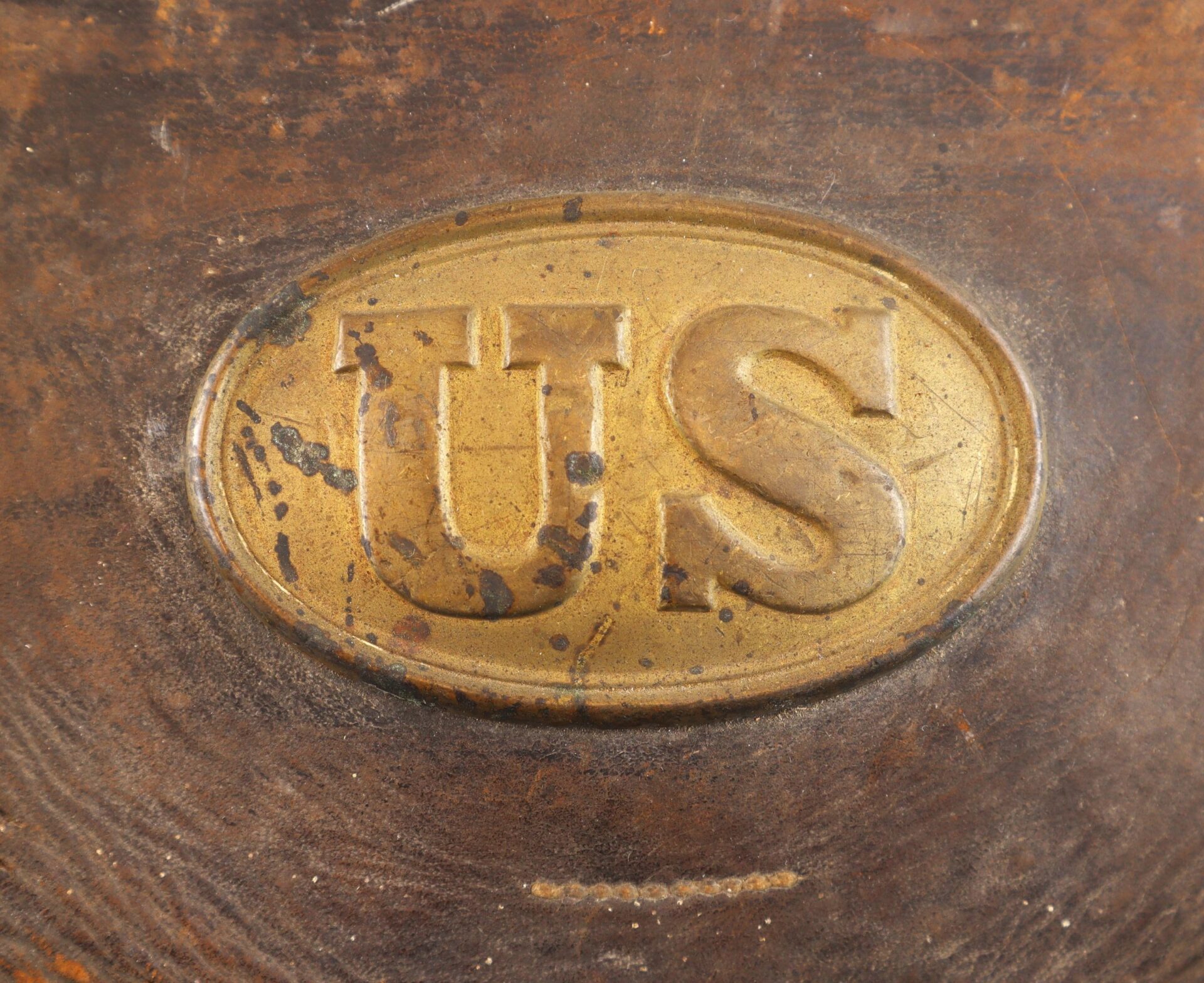 Complete Cartridge Box / SOLD: Oval brass plate with raised "US" letters, featured on a worn brown background showing scratches and tarnish.