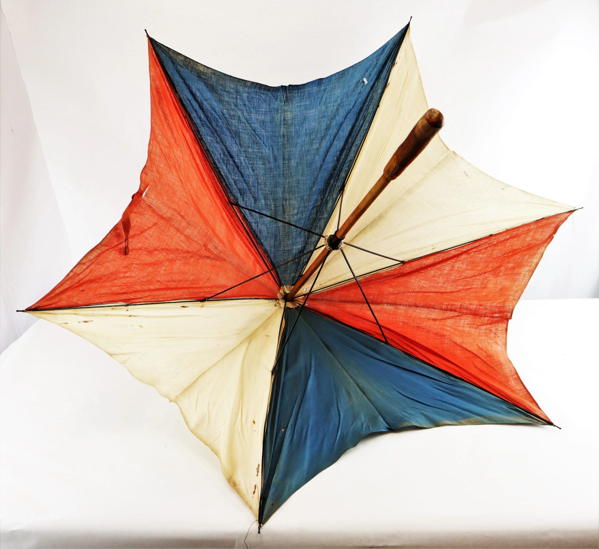 The Patriotic Parasol, a vintage umbrella with alternating blue, red, and cream panels, is open on a white surface; its fabric shows signs of wear and staining.