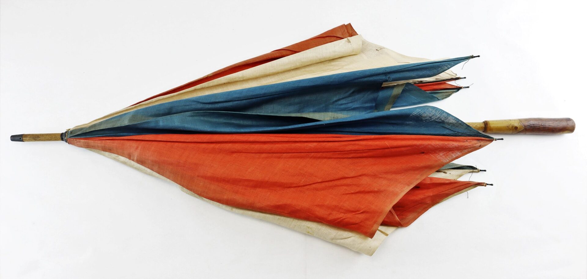 The Patriotic Parasol, a closed vintage umbrella with red, blue, and beige panels and a wooden handle showing signs of wear, rests on a white background.