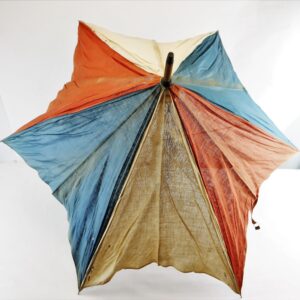 A top-down view of the Patriotic Parasol, an old, worn umbrella featuring blue, red, and beige multicolored fabric panels set against a white background.