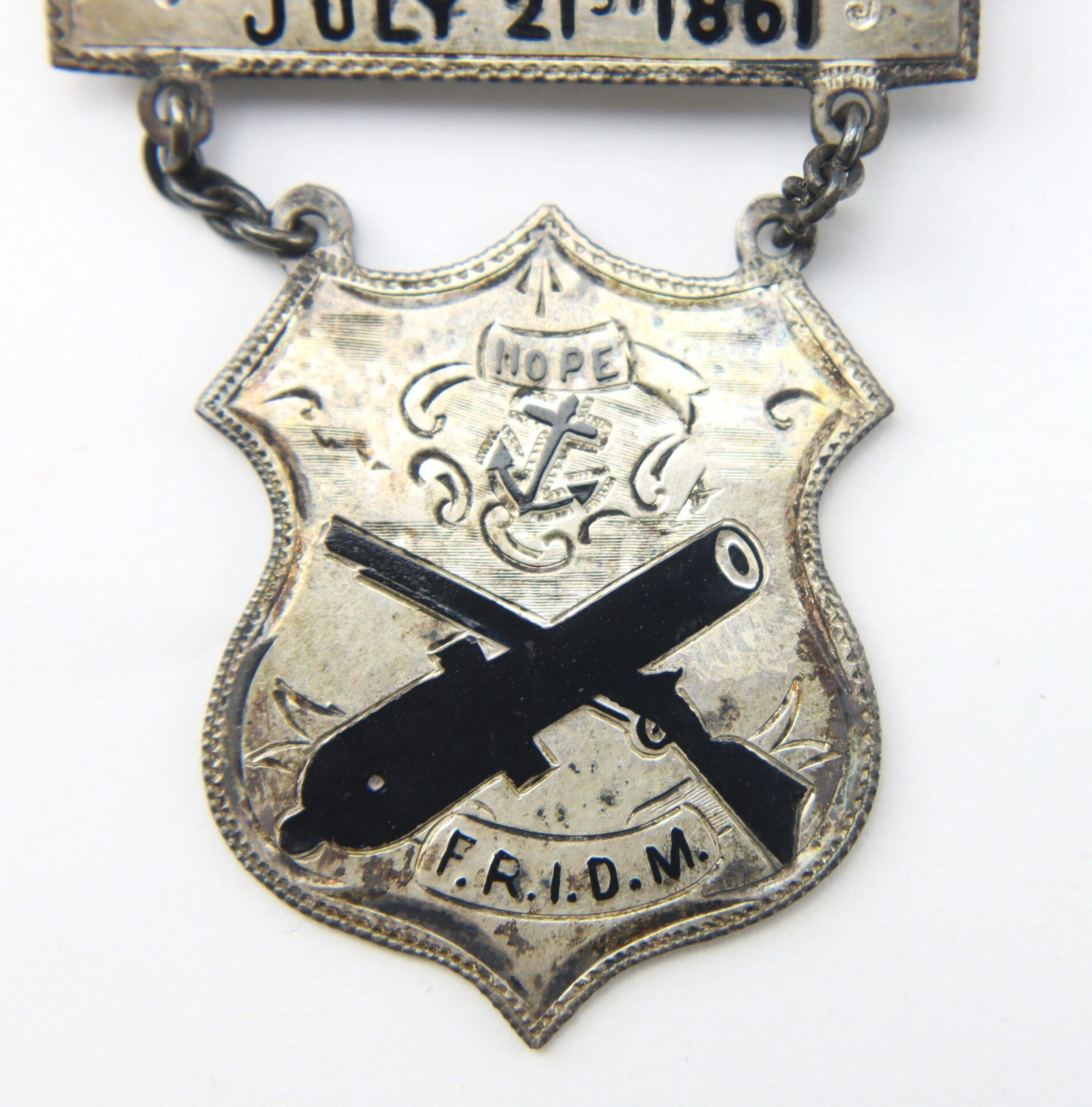 The Rhode Island Regimental Badge “Bull Run July 21 1861” F.R.I.D.M. for the First Rhode Island Detached Militia is a silver shield-shaped medal with black crossed weapons, an anchor with "HOPE," and "F.R.I.D.M." engraved at the bottom.
