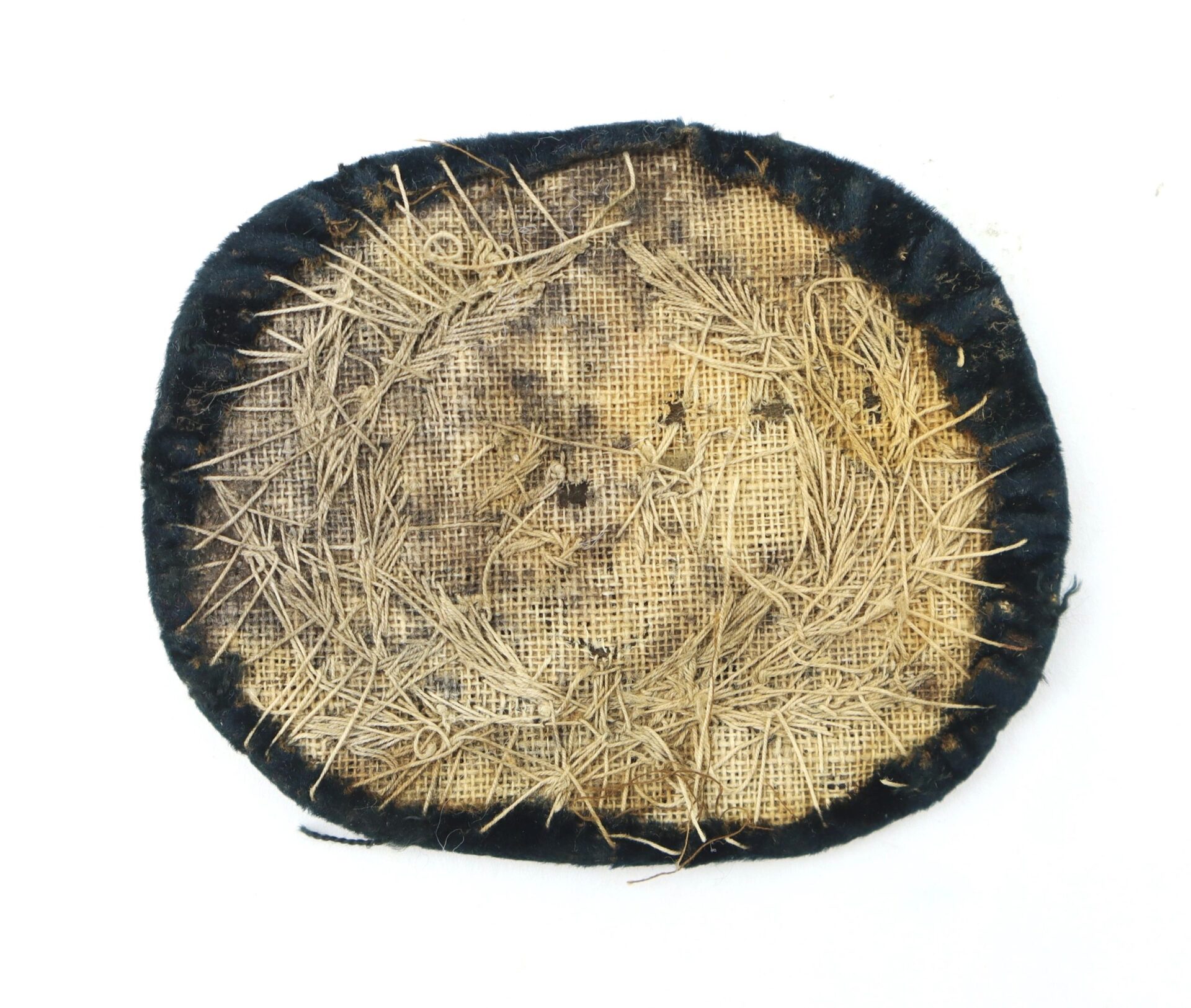 A round, worn fabric pad with a black border and coarse beige center, featuring visible stitching and scattered pine needles, resembling an embroidered U.S. Officer's Hat Insignia.