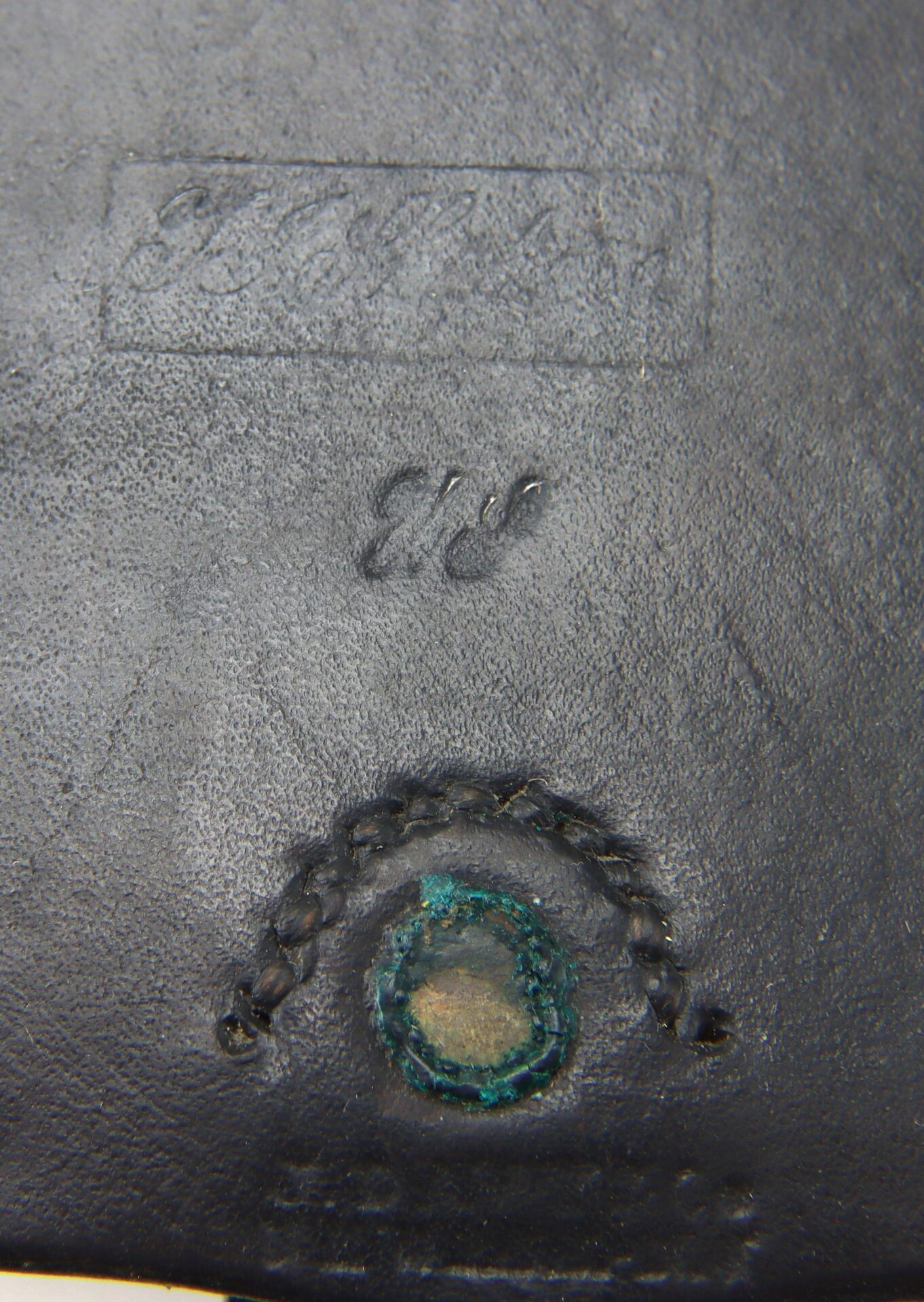 Close-up of the Pistol Cartridge Box / SOLD, showing black leather with embossed and stamped markings, visible stitching, and a circular greenish corrosion near the bottom center.