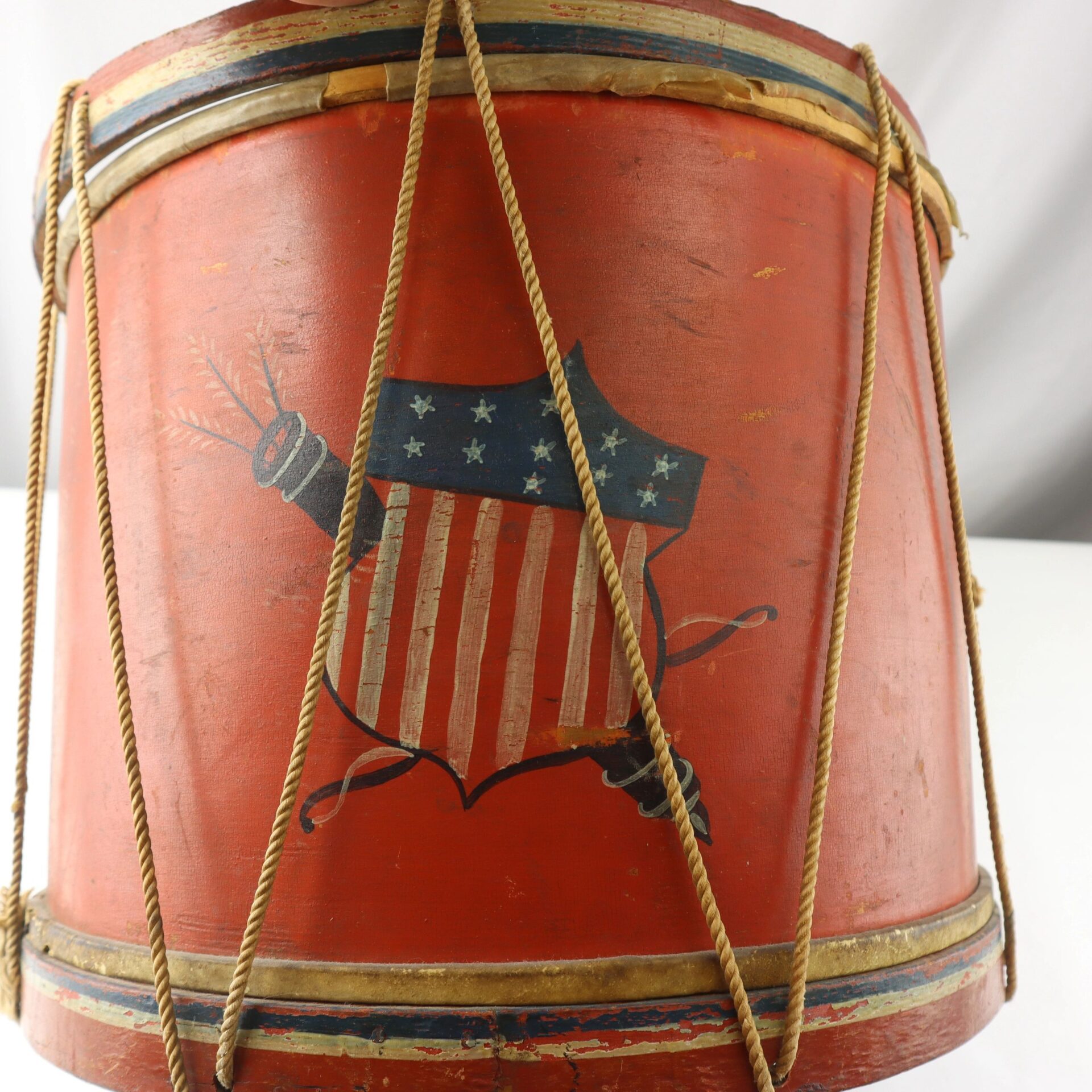 The Civil War Painted Drum is a vintage red drum with rope tensioning, decorated with an American shield featuring stars, stripes, and crossed cannon ramrods.