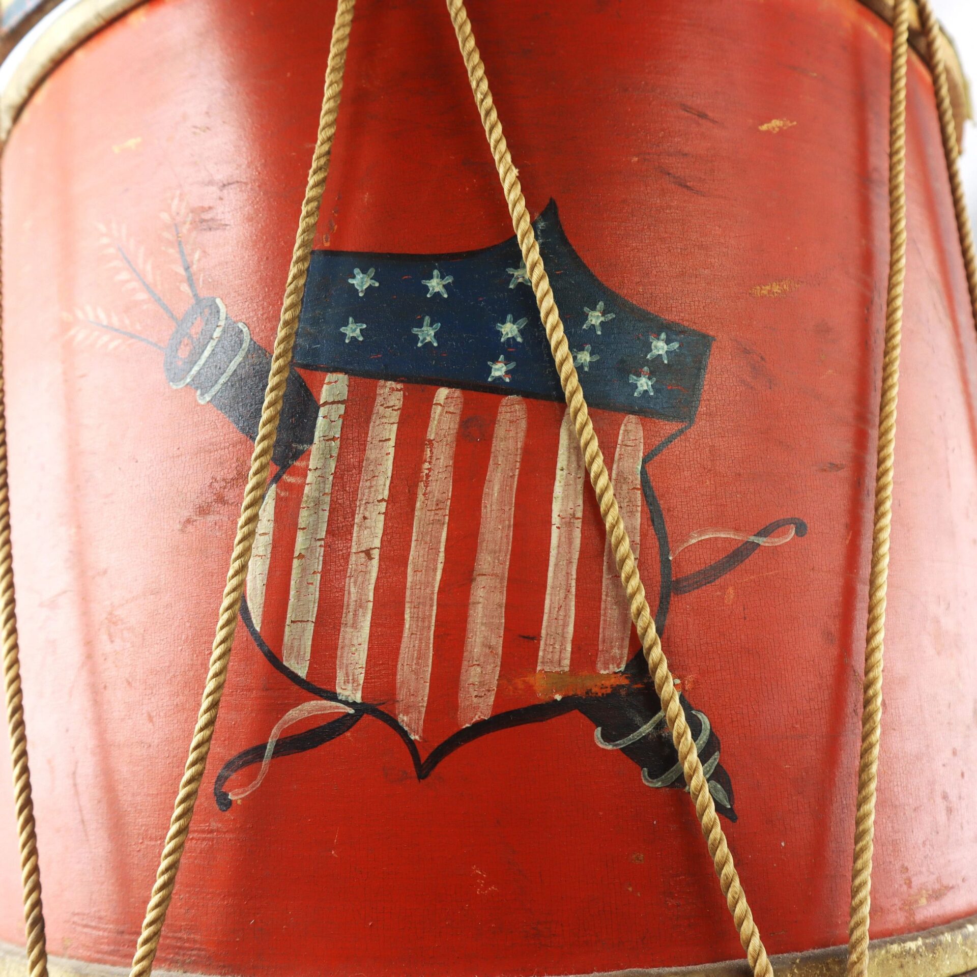 A close-up of the Civil War Painted Drum, showcasing a red body with a shield featuring stars and stripes like the American flag, and visible rope tension cords.
