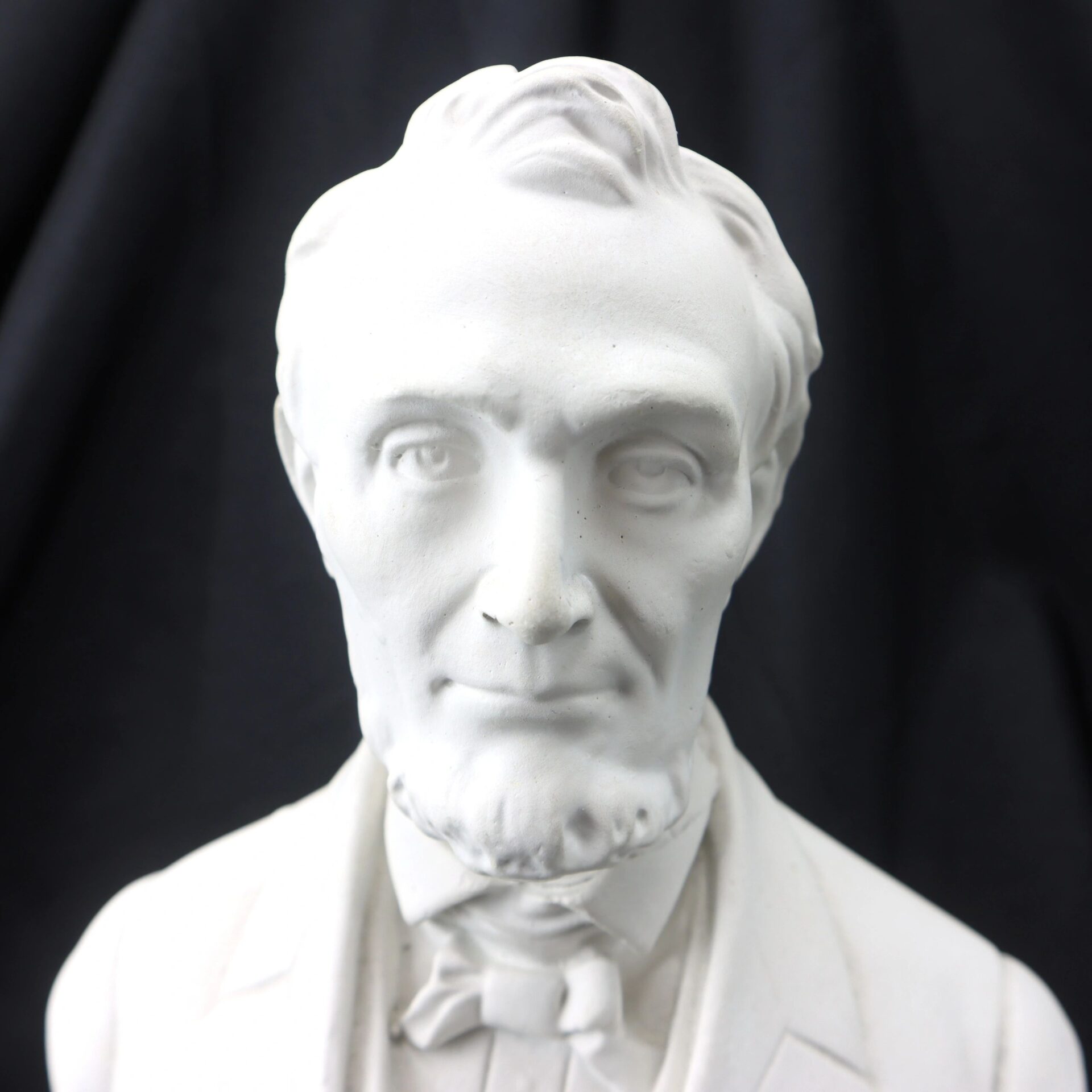White marble bust of Abraham Lincoln with a beard and bow tie, set against a plain black background.