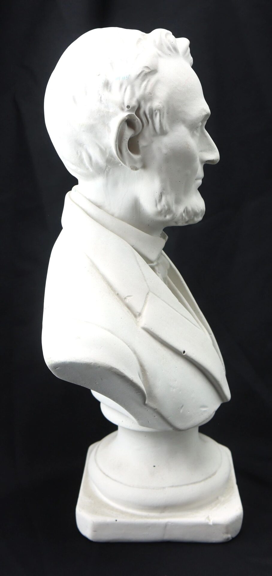 White plaster bust of Abraham Lincoln, depicted in profile facing right in a suit, set against a plain black background.