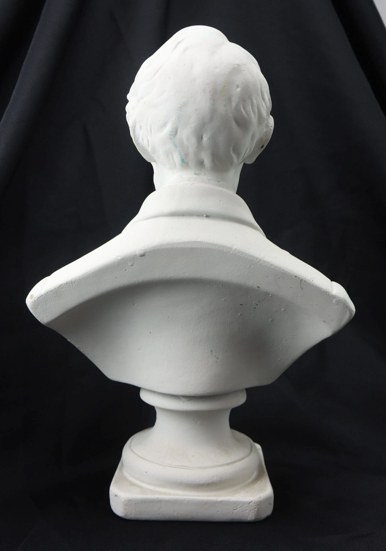 A white plaster bust sculpture of Abraham Lincoln viewed from the back, set against a black background.