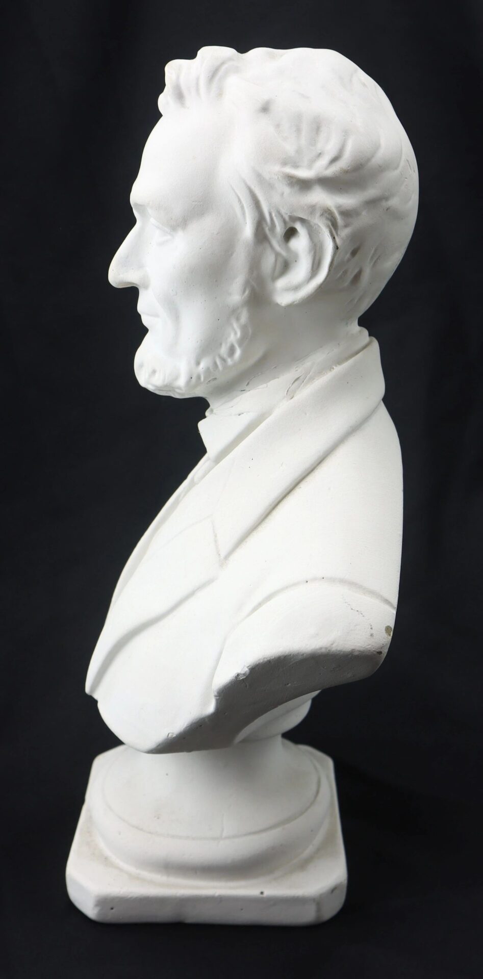 A white plaster bust of Abraham Lincoln, depicted in profile with a beard and suit jacket, set against a black background.