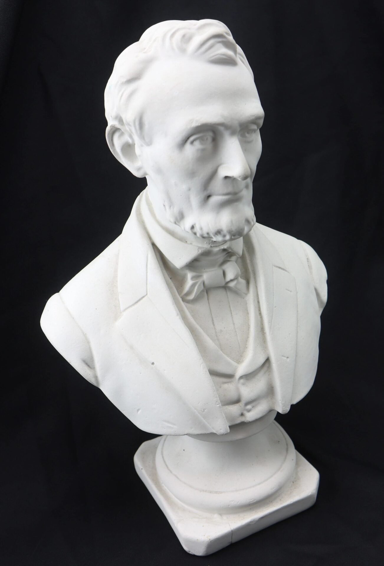 White marble bust of Abraham Lincoln, depicted with a beard and wearing a suit and bow tie, set against a black background.