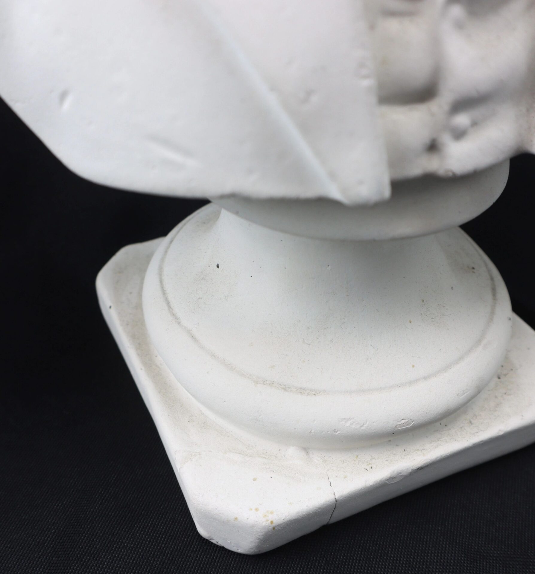 Close-up of the base of the "Abraham Lincoln" white plaster bust on a black background, highlighting the square pedestal and part of the lower sculpture.