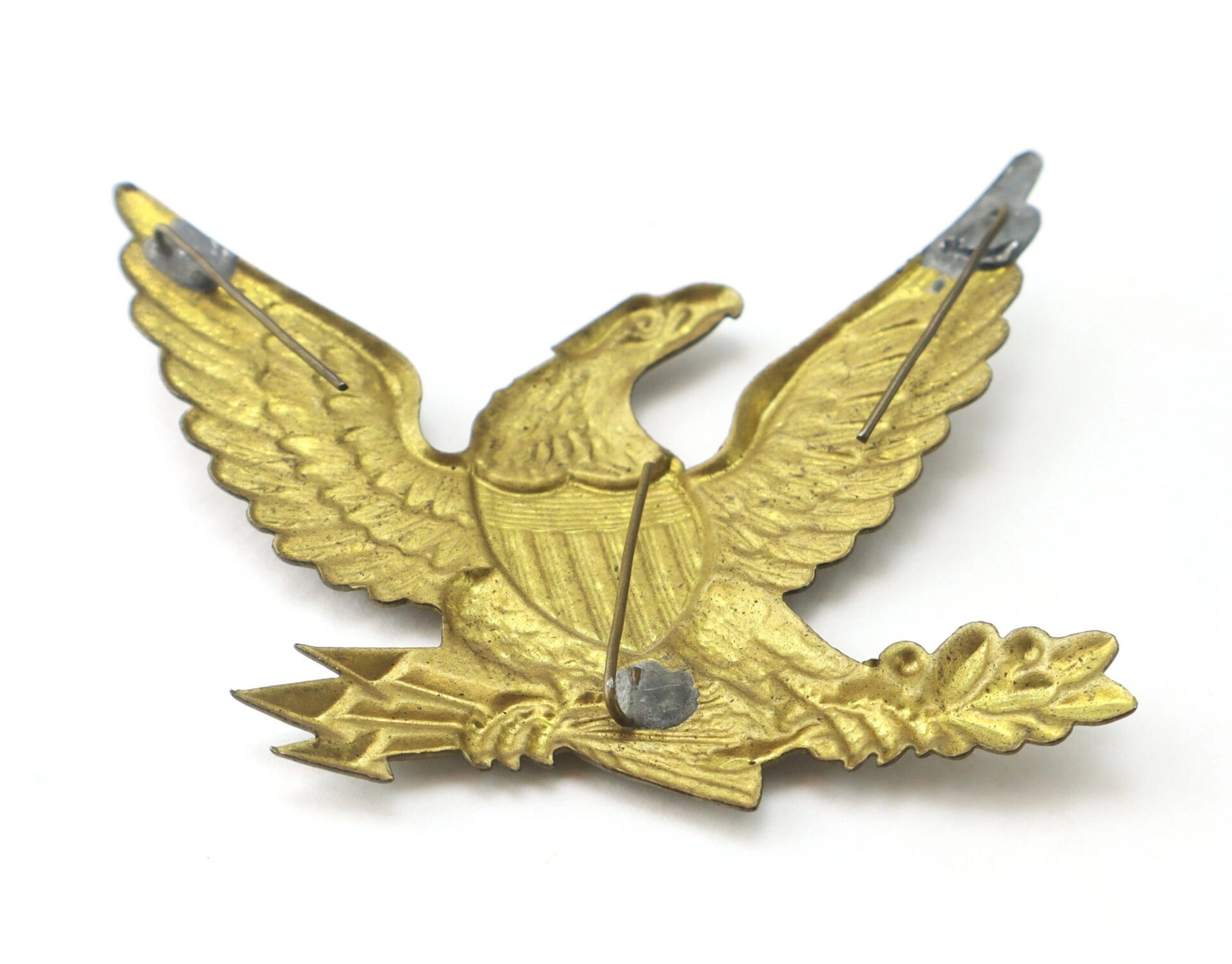 The Eagle Hat Device is a gold-colored metal emblem of an eagle with outstretched wings, clutching arrows and an olive branch; it features pin fastenings on the back.