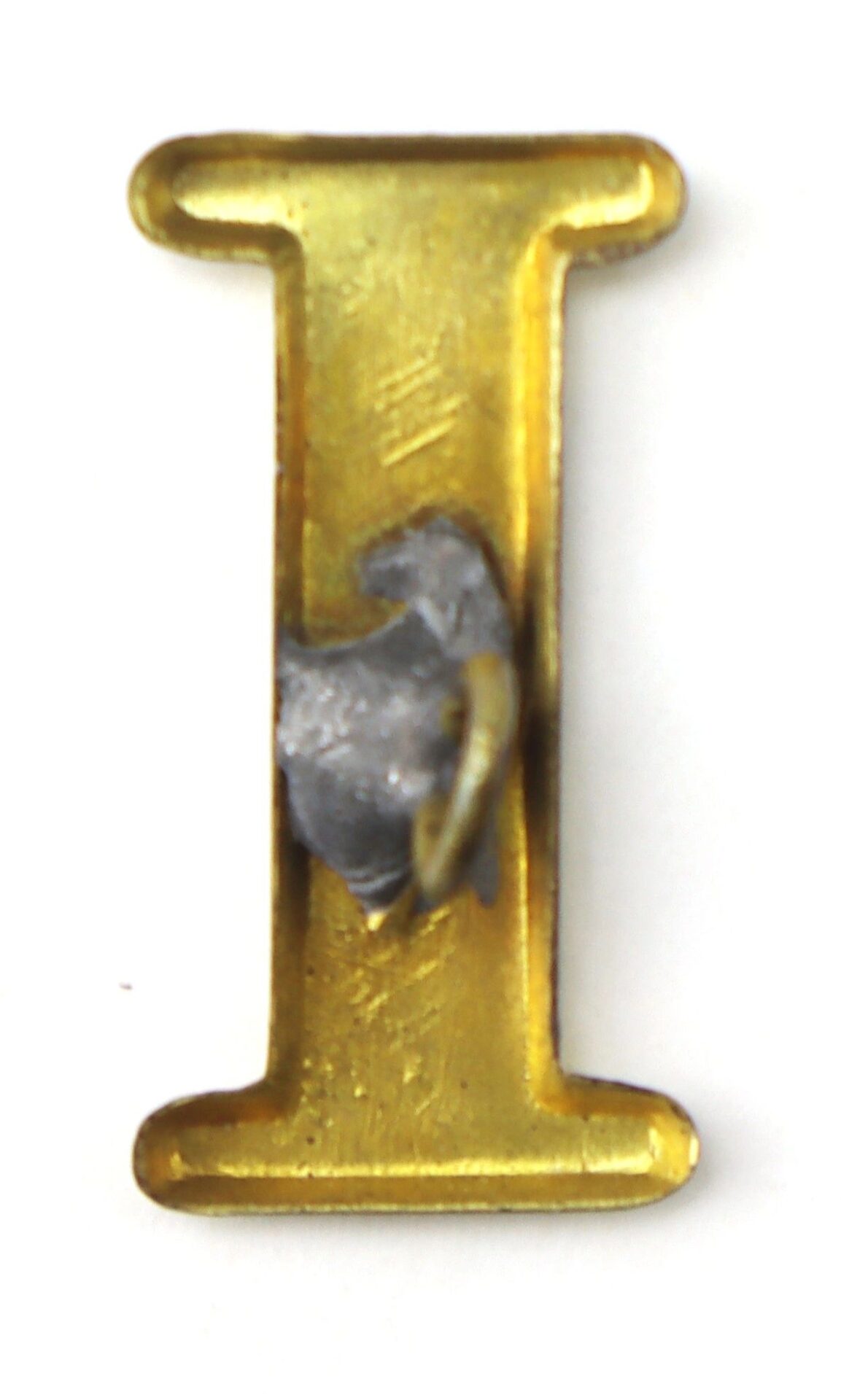 The Regimental Company Letter I is a gold metal letter "I" featuring a gray, irregularly-shaped blob attached at its center.