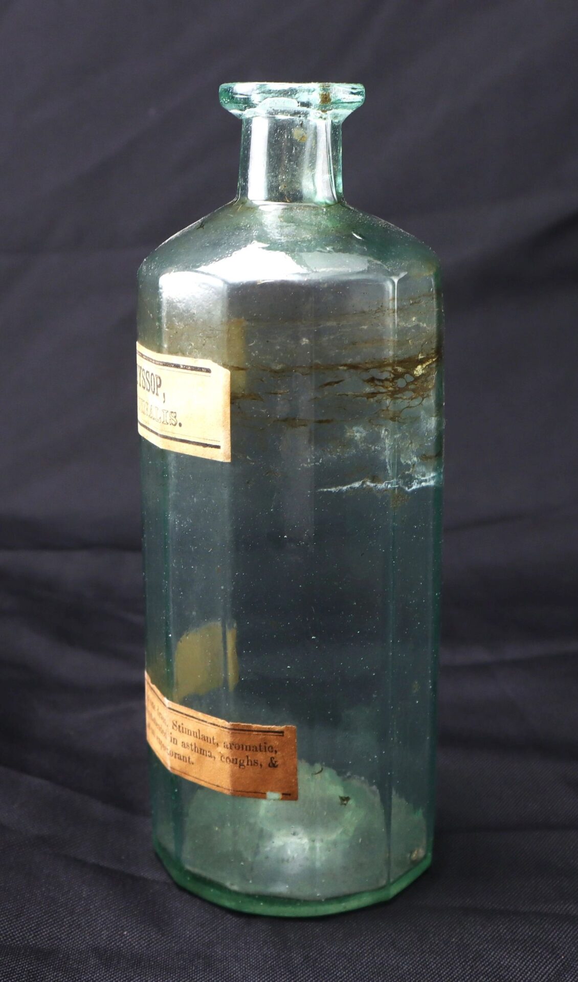 A vintage Apothecary Bottle – Fluid Extract Hyssop, featuring a narrow neck and original paper labels, is partially filled with cloudy liquid and displayed against a dark background.