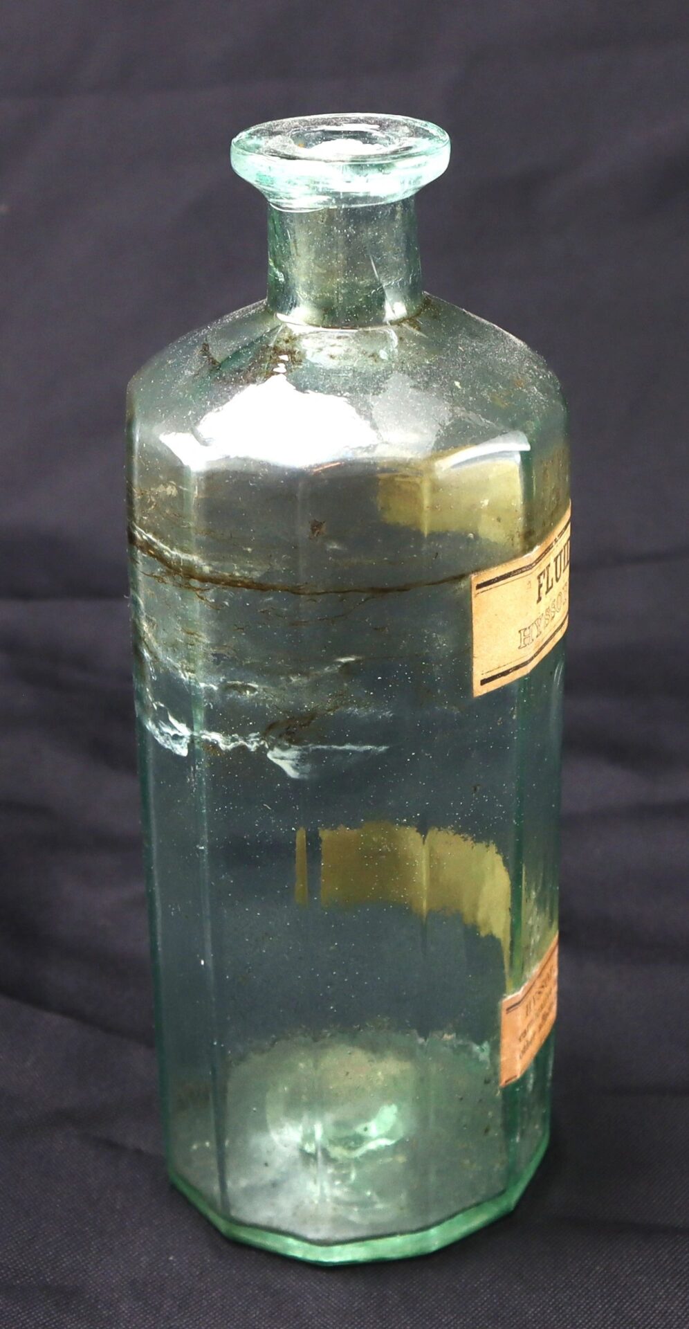 A vintage, clear glass Apothecary Bottle – Fluid Extract Hyssop with an octagonal shape, partial paper labels, and some residue inside is set against a dark background.