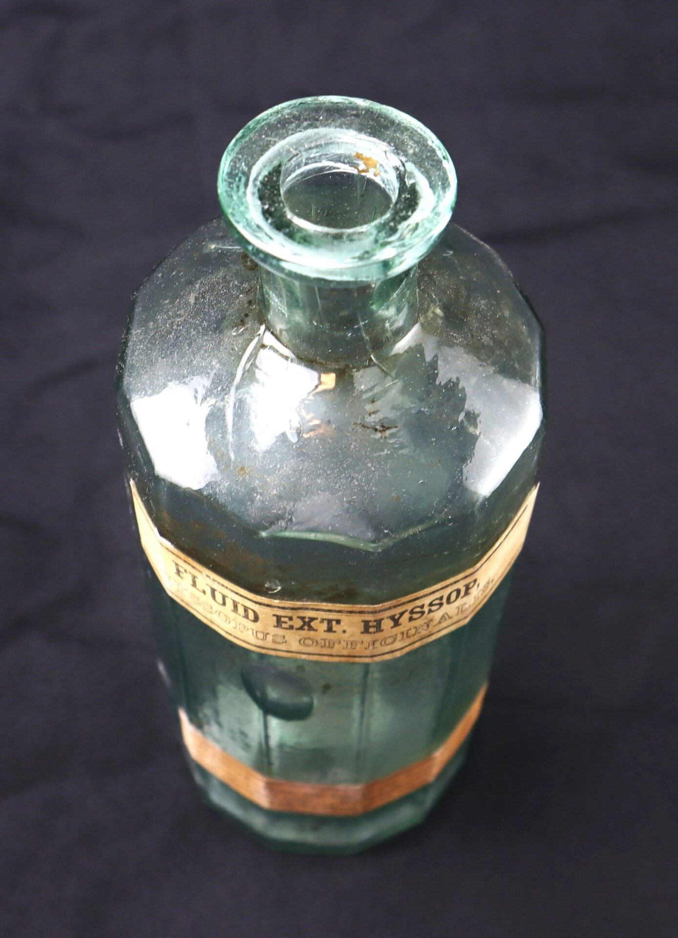 The Apothecary Bottle – Fluid Extract Hyssop is a vintage clear glass bottle with a narrow neck and a "FLUID EXT. HYSSOP" label, displayed against a black background.