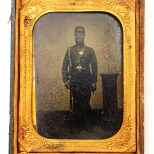 A vintage photograph of a man in a Civil War-era military uniform standing beside a podium with a small American flag, framed in an ornate gold case.
