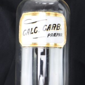 A clear glass apothecary bottle with a stopper, labeled "CALC. CARB. PREPAR." against a black background.