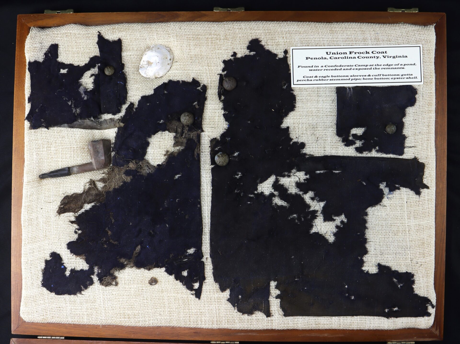 Display case showing heavily deteriorated fragments of a dark fabric frock coat, two buttons, a pipe, and a descriptive plaque about the coat’s origin from a Union soldier in Virginia.