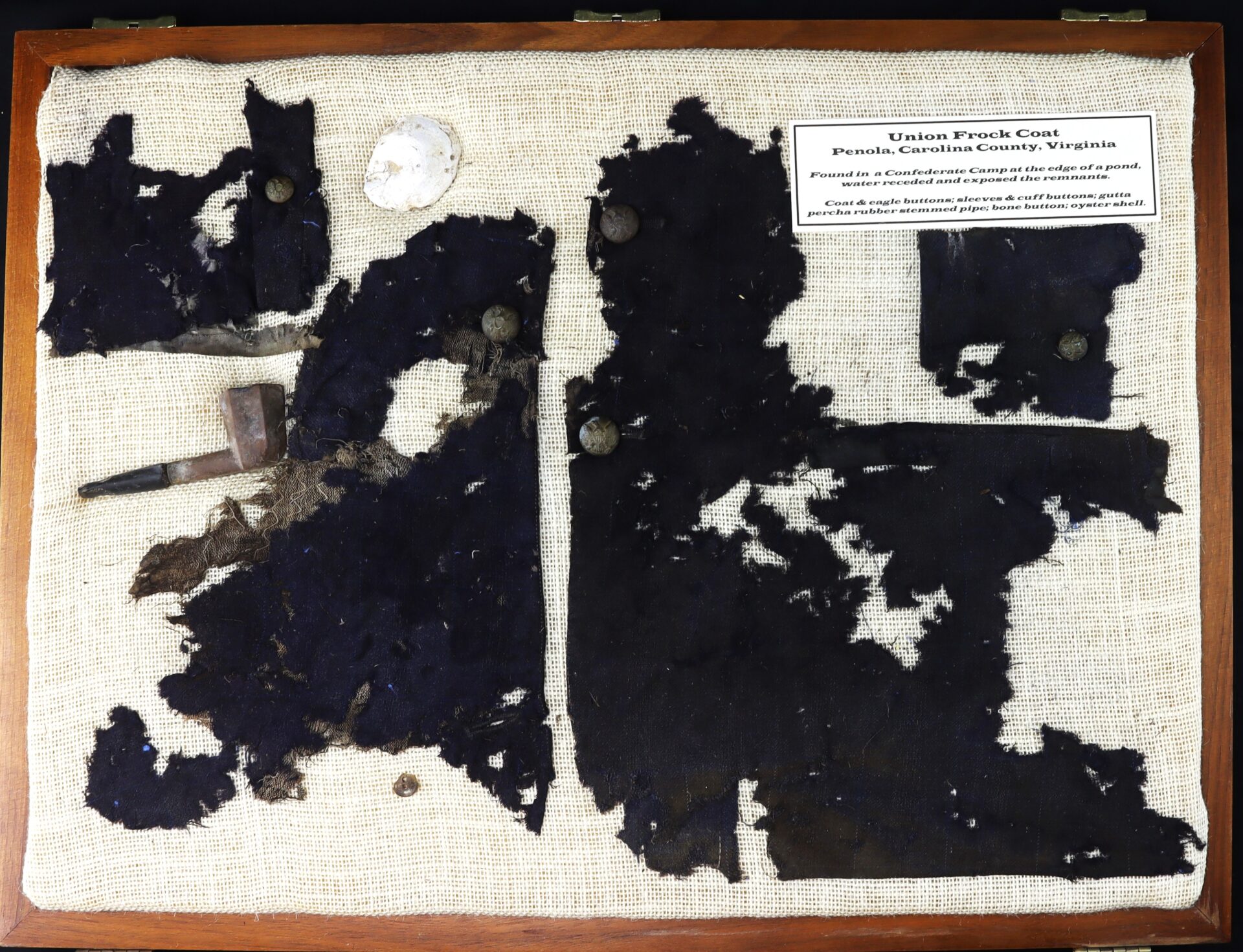Display case containing fragments of a dark fabric Union frock coat, a pipe, four buttons, a white disc, and an informational label about their Civil War origin from Panola, Virginia.