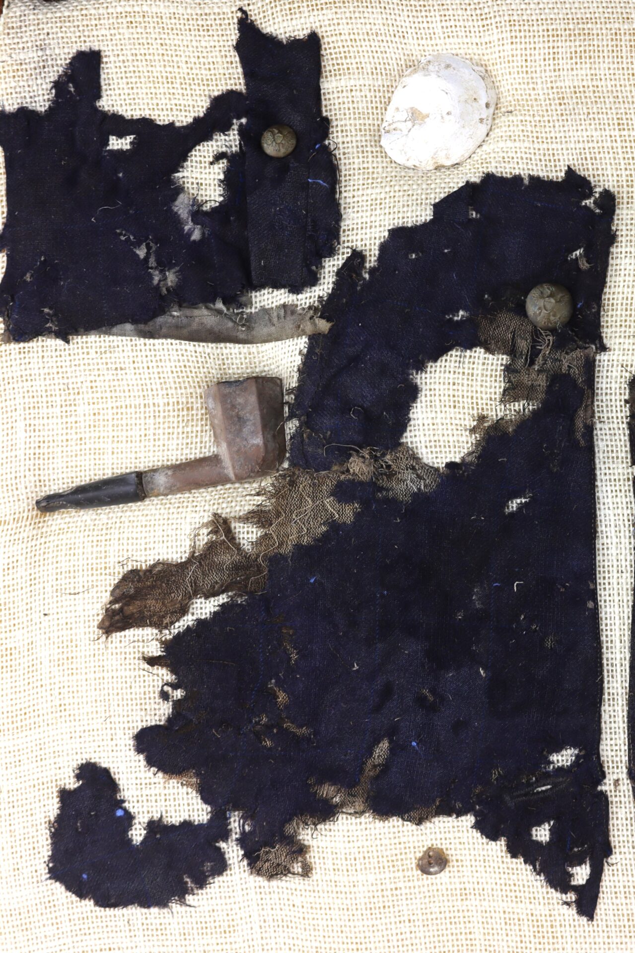 Dark, tattered fabric fragments with metal buttons, a corroded pipe, and a round white object arranged on a coarse, light-colored cloth background.