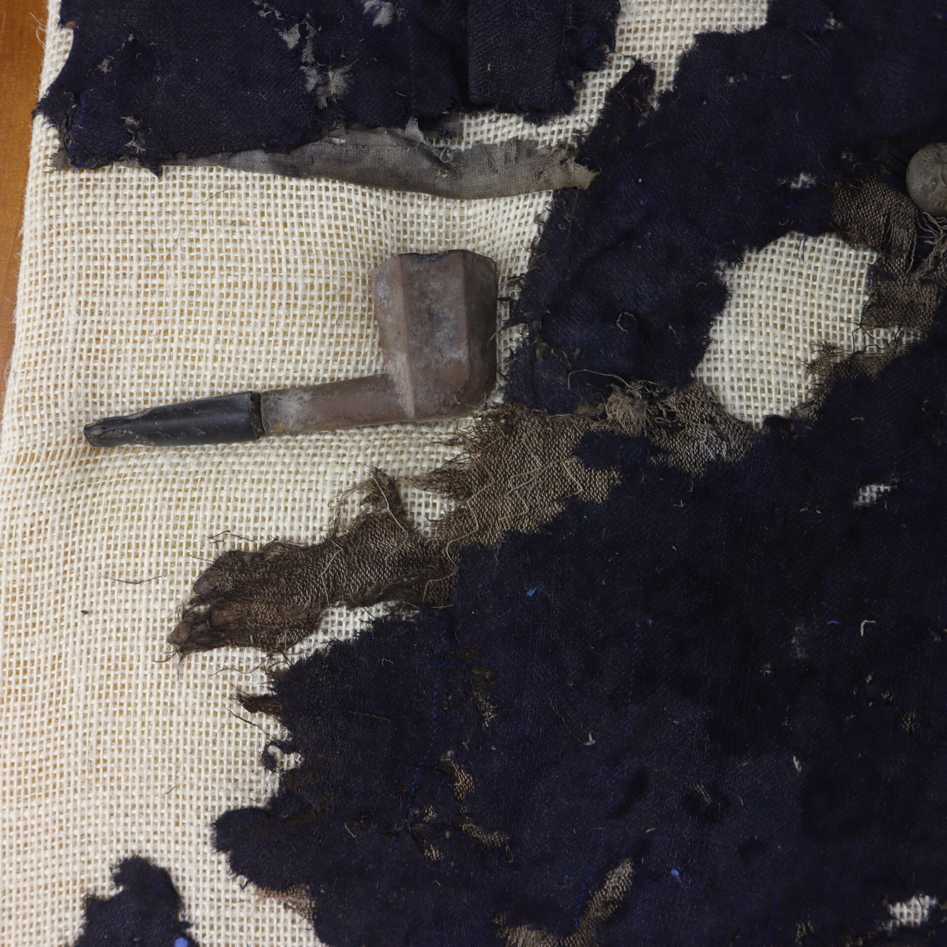 A rusted metal pipe lies on top of frayed dark fabric and a cream-colored woven textile background.