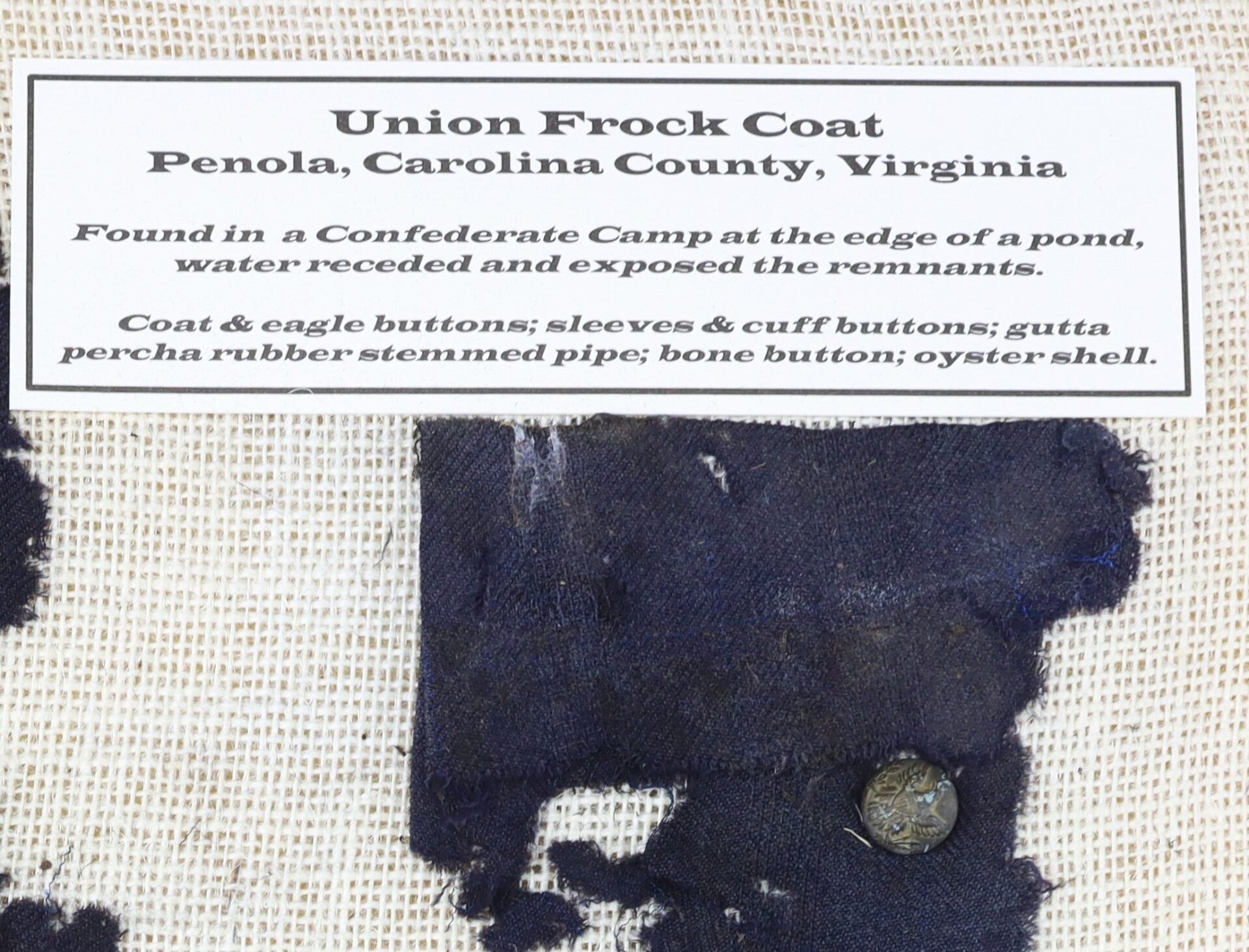 A worn fragment of a Union frock coat with a button, displayed with a label describing its discovery in a Confederate camp in Penola, Virginia, and listing found artifacts.