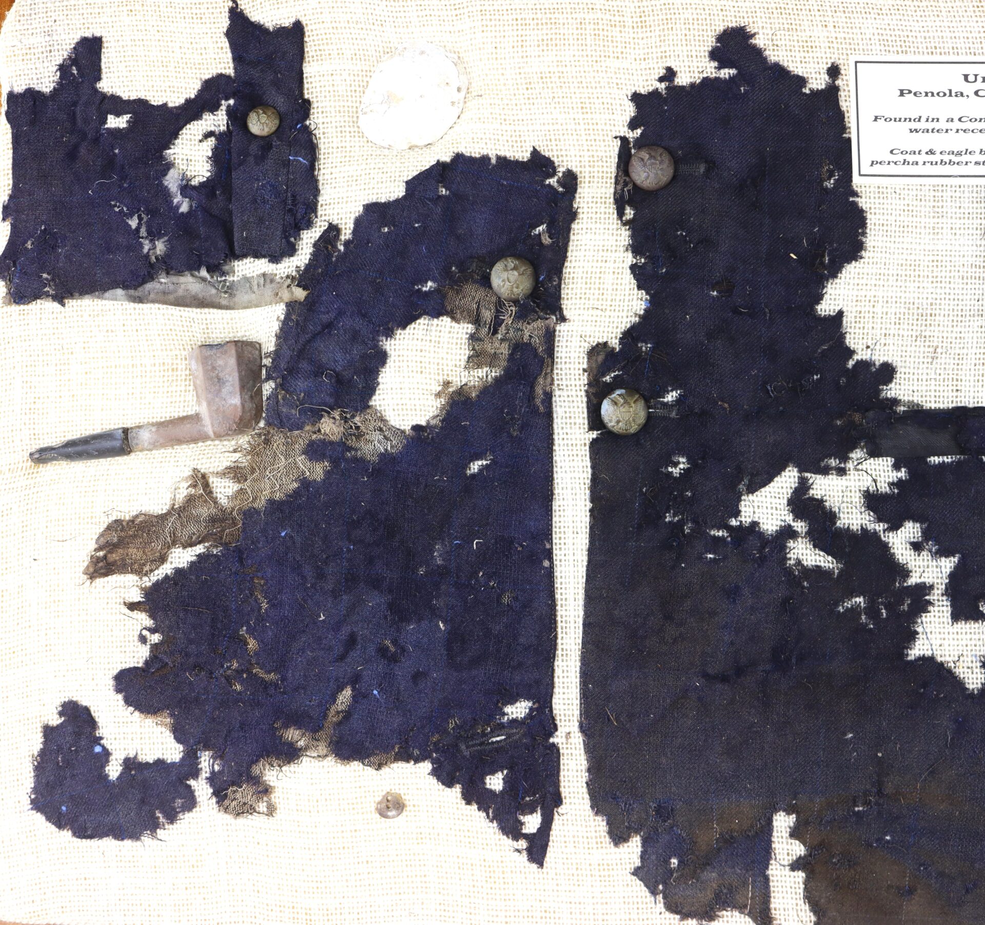 Fragments of dark blue fabric with buttons, a white coin, and a small pipe displayed on a light background with a label in the upper right corner.