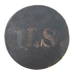 A round, weathered, black surface with the letters "MS" and "H" scratched into it, along with other faint markings.