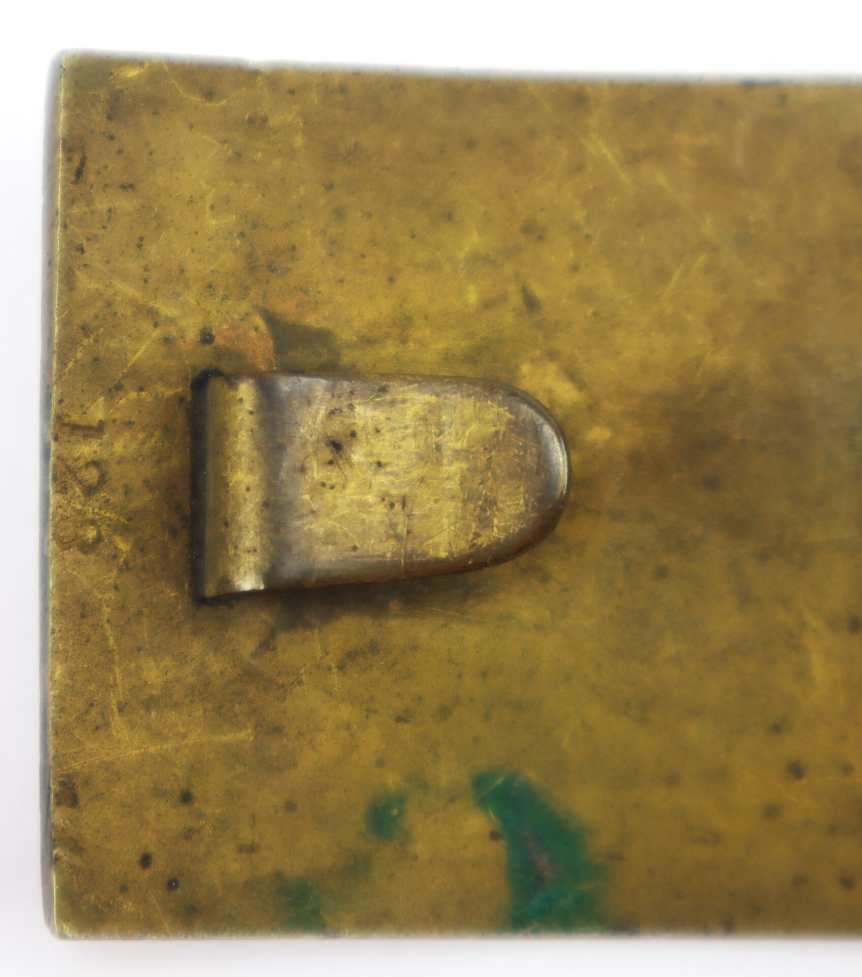 Close-up of a tarnished, rectangular brass object with a curved metal clip attached to its surface.