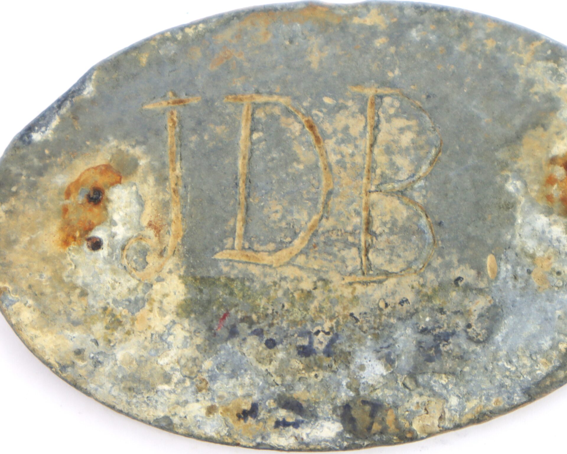 Oval-shaped, weathered metal tag with the engraved initials "JDB"; surface shows rust spots and corrosion.