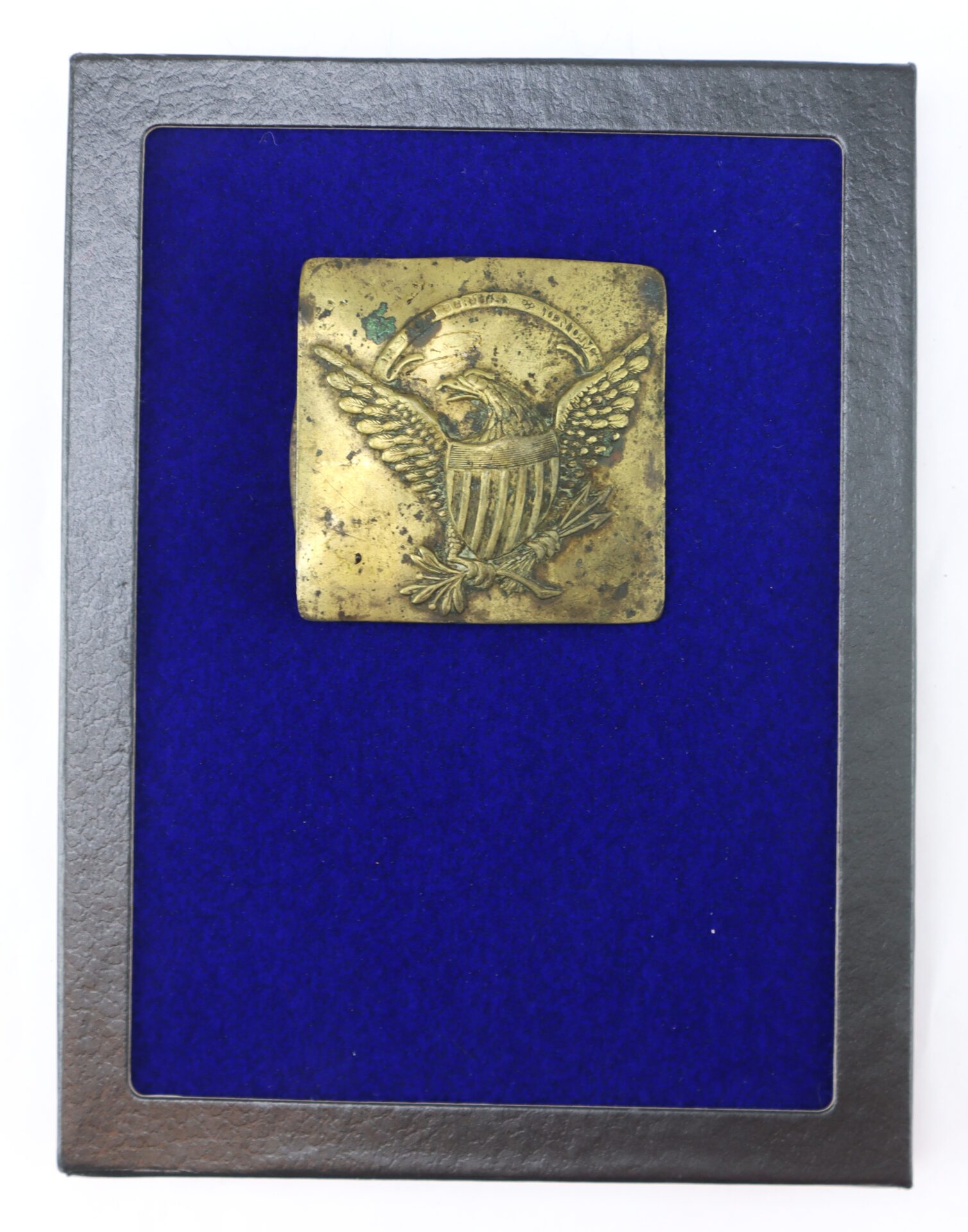 A brass military belt plate featuring an eagle emblem is displayed in a black frame with a blue velvet background.