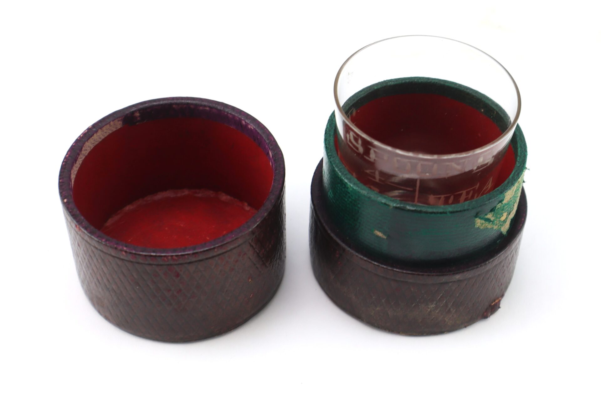 A small, round, open container with a red interior holds a glass vessel filled with dark liquid beside its empty matching lid.