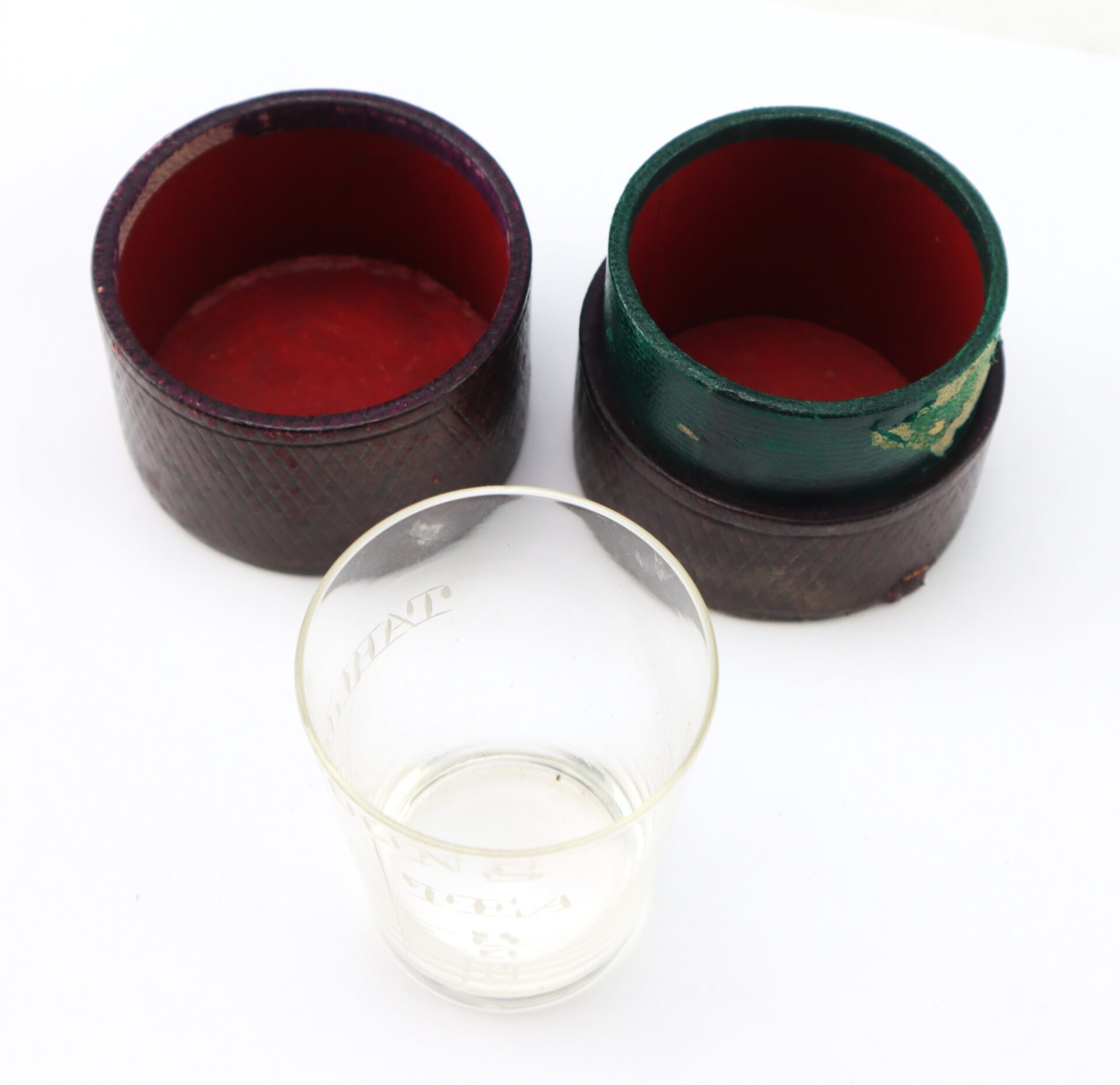 A small clear glass sits in front of two open, round, red-lined cases—one dark brown, one green—on a white surface.