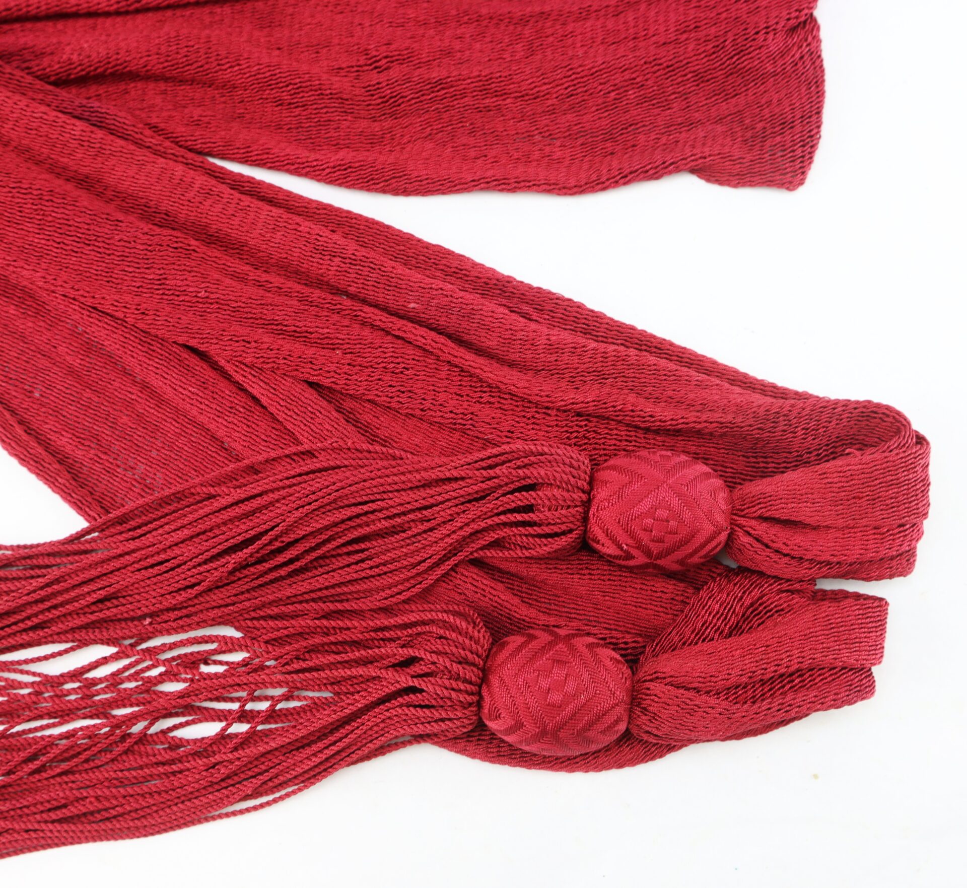 Close-up of two red woven tassels with textured strands and decorative knotted balls at the ends, laid out on a white background.