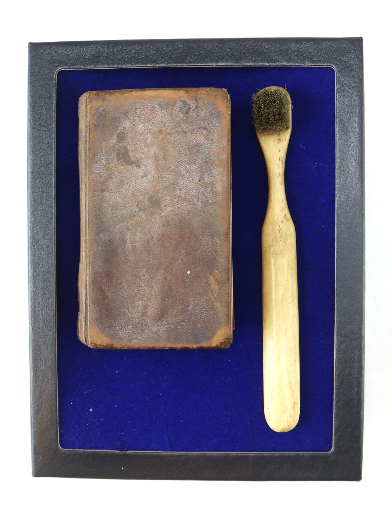 A Confederate Bible with a worn brown leather cover sits beside an old wooden toothbrush on blue velvet, all displayed inside a black frame.