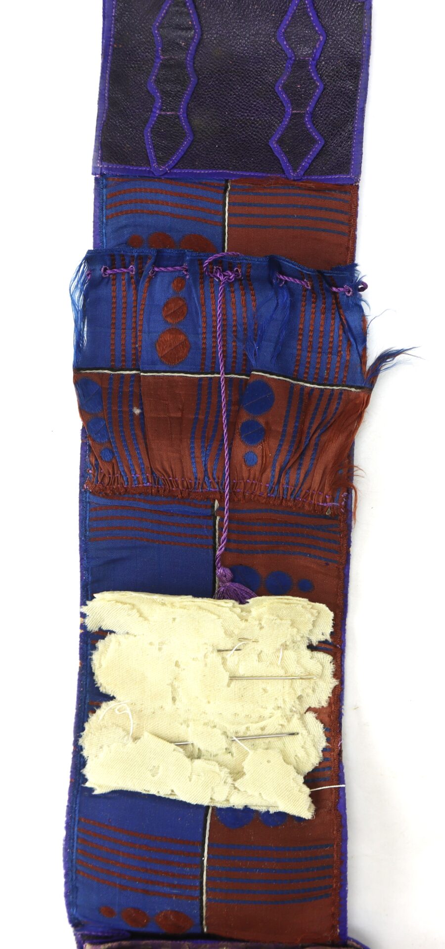 A narrow textile strip with geometric patterns in blue, red, and purple, frayed edges, a purple wavy design, and a patch of beige fabric sewn near the bottom.